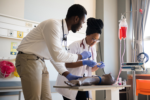 Young male and female doctors practicing IV Drip on artificial limb on desk. Healthcare colleagues are in training class. They are in hospital.